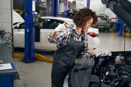 Téléchargez les photos : Multiracial jeune réparateur automatique vérifie le niveau d'huile dans la voiture, la femme travaille dans des combinaisons de travail confortables - en image libre de droit