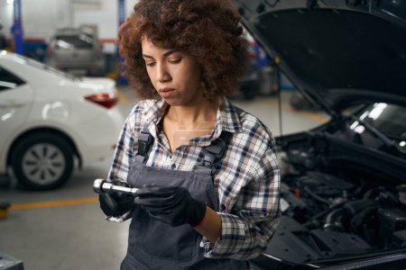 Téléchargez les photos : Employé d'atelier de réparation automobile multiracial en vêtements de travail debout près de la voiture, femme tenant le détail - en image libre de droit