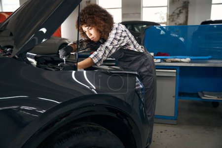 Téléchargez les photos : Jeune réparateur multiracial en chemise à carreaux travaille sous le capot de la voiture, une femme est dans un atelier de réparation automobile - en image libre de droit