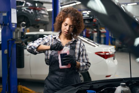 Foto de Bastante empleado de un taller de reparación de automóviles sostiene contenedor con una lavadora de vidrio en sus manos, mujer en guantes de trabajo - Imagen libre de derechos