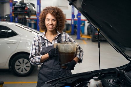 Foto de Sonriendo joven mecánico de automóviles se encuentra cerca de coche con un contenedor de aceite de coche, mujer trabaja en taller de reparación de automóviles modernos - Imagen libre de derechos