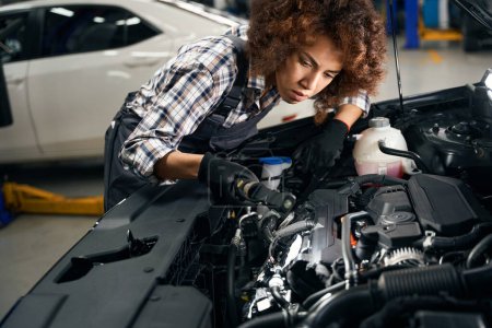Foto de Empleado de un taller de reparación de automóviles inspecciona un coche bajo el capó, una mujer trabaja en ropa de trabajo cómoda - Imagen libre de derechos