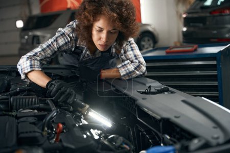 Téléchargez les photos : Jeune réparateur multiracial inspecte le moteur sous le capot de la voiture, la femme est dans l'atelier de réparation automobile - en image libre de droit