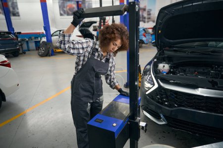 Photo for Young repairwoman uses special equipment for service maintenance of the car, the car stands next to the open hood - Royalty Free Image
