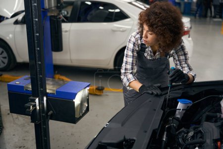 Photo for Young female employee of car repair shop uses modern equipment to repair a car, car is standing with open hood - Royalty Free Image