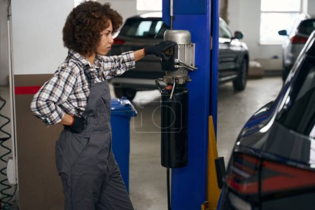Téléchargez les photos : Mécanicien féminin soulève la voiture sur un ascenseur spécial pour l'inspection, une femme en salopette de travail - en image libre de droit