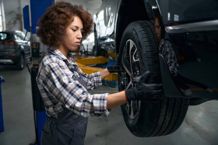 Téléchargez les photos : Femme mécanicien automobile examine attentivement une roue de voiture, une voiture sur un service régulier dans un atelier de réparation automobile moderne - en image libre de droit