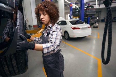 Téléchargez les photos : Jeune réparatrice bouclée inspecte une roue de voiture, la voiture est entretenue dans un atelier de réparation automobile - en image libre de droit