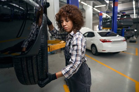 Téléchargez les photos : Femme réparateur multiracial inspectant une roue de voiture, la voiture est entretenue dans un atelier de réparation automobile - en image libre de droit
