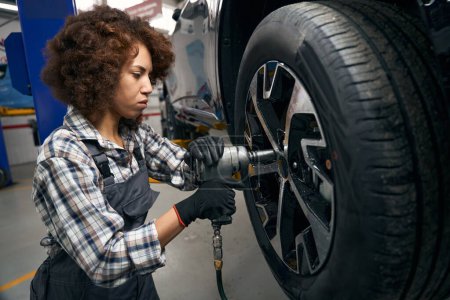 Téléchargez les photos : Femme réparateur utilise un tournevis pneumatique pour travailler avec une roue de voiture, femelle en salopette de travail - en image libre de droit
