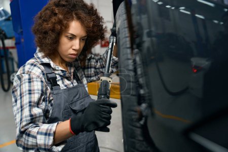 Téléchargez les photos : Jeune réparateur automobile en vêtements de travail regarde la roue d'une voiture, une femme travaille dans un atelier moderne - en image libre de droit