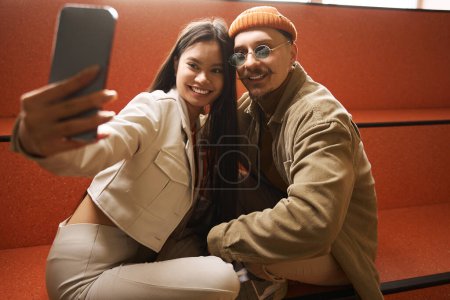 Foto de Sonriendo feliz elegante joven asiática mujer y caucásico hombre tomando selfie en banco - Imagen libre de derechos
