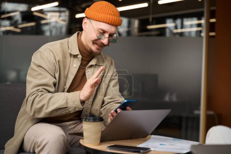 Foto de Sonriente empleado de la compañía sentado en el portátil saludando a alguien en la pantalla táctil del teléfono inteligente - Imagen libre de derechos