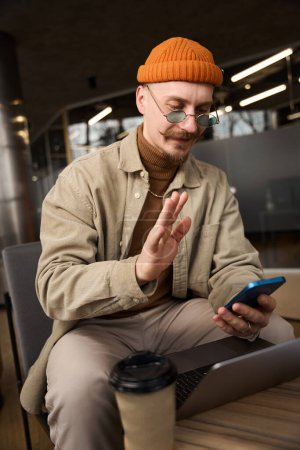 Téléchargez les photos : Employé de bureau agréable assis à un ordinateur portable agitant quelqu'un sur smartphone - en image libre de droit