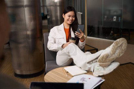 Téléchargez les photos : Employé souriant de l'entreprise avec tasse en papier dans les mains assis dans la chaise avec les pieds sur le bureau en regardant l'écran tactile du smartphone - en image libre de droit