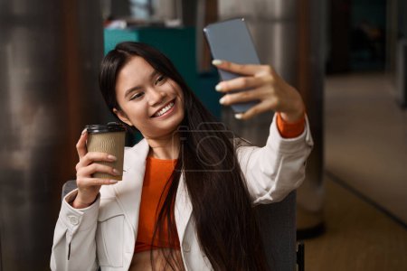 Téléchargez les photos : Portrait taille haute de joyeuse employée d'entreprise avec tasse en papier à la main se photographiant avec un appareil photo smartphone - en image libre de droit