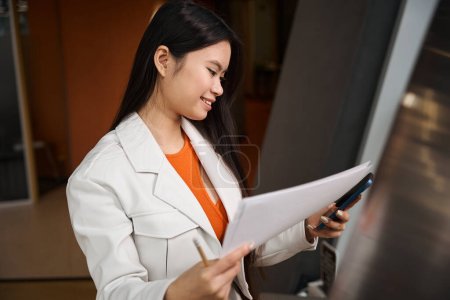 Photo for Smiling female office worker with documents in hand looking at smartphone touchscreen - Royalty Free Image