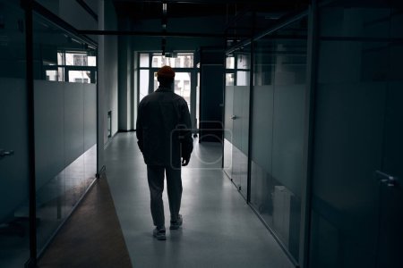 Photo for Back view of company employee walking along dark corridor in office building - Royalty Free Image