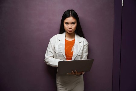 Photo for Serious stylish young woman with portable computer standing against wall and looking before her - Royalty Free Image