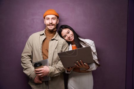 Photo for Smiling happy Asian woman and pleased Caucasian man with laptops standing by wall - Royalty Free Image