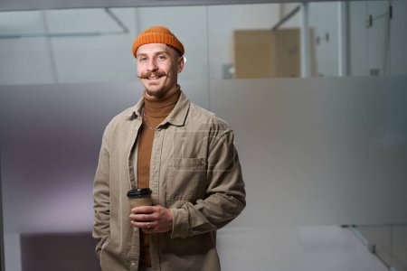 Foto de Retrato en la cintura del empleado alegre de la empresa con taza de papel en las manos de pie solo en el pasillo de la oficina - Imagen libre de derechos