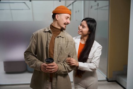 Smiling joyous man with paper cup in hands looking at his happy female companion in office corridor