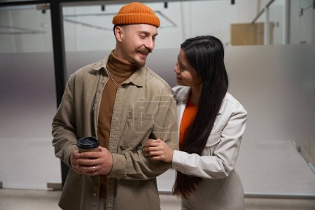 Téléchargez les photos : Homme souriant avec tasse en papier dans les mains regardant sa joyeuse compagne dans le couloir du bureau - en image libre de droit