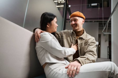 Photo for Smiling happy young man and woman seated on sofa hugging each other in office corridor - Royalty Free Image