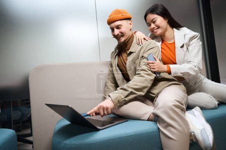 Téléchargez les photos : Joyeux gars pointant l'index à l'écran de l'ordinateur portable pour sourire femelle assise à côté de lui sur le canapé - en image libre de droit