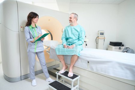 Téléchargez les photos : Heureux patient masculin remplissant un questionnaire avant le diagnostic IRM tout en regardant le médecin féminin - en image libre de droit