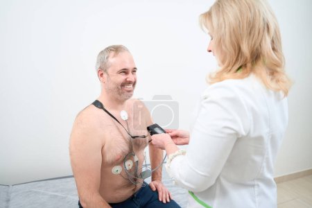 Photo for Doctor using holter monitor device and daily blood pressure recorder for male patient in clinic - Royalty Free Image