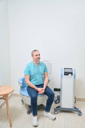 Téléchargez les photos : Sourire patient homme caucasien attendant un médecin assis sur la salle d'examen - en image libre de droit