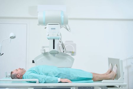 Téléchargez les photos : Homme adulte allongé sur un lit et attendant une machine à rayons X pour scanner à l'hôpital. Hôpital moderne avec un équipement technologiquement avancé - en image libre de droit