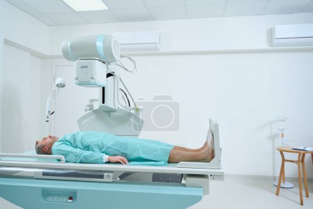 Foto de Hombre adulto acostado en la cama esperando a que la máquina de rayos X escanee en busca de lesiones en la clínica moderna - Imagen libre de derechos