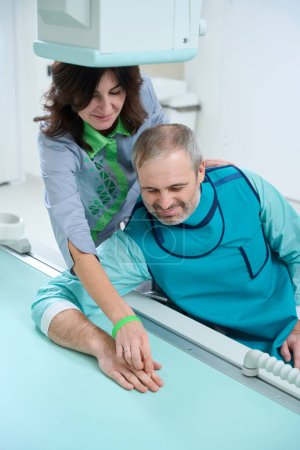 Photo for Smiling female doctor correcting patient hand while helping to making x-ray on modern equipment - Royalty Free Image