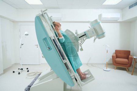 Photo for Side view of adult man in a medical gown taking radiograph an X-Ray machine to scan for injury in clinic - Royalty Free Image