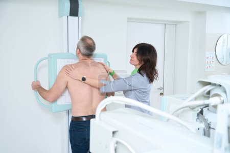 Téléchargez les photos : Homme adulte en blouse médicale soumis à une radiographie pulmonaire, médecin femme aidant pour lui - en image libre de droit