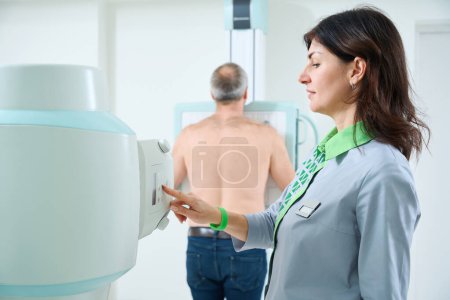 Foto de Radióloga sonriente que toma rayos X del hombre mientras opera la máquina de rayos X - Imagen libre de derechos