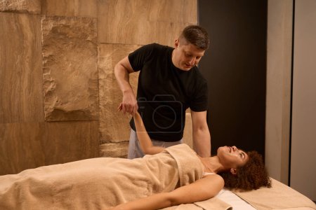 Téléchargez les photos : Femme joyeuse visiteur couché sur le bureau de massage et relaxant pendant le massage des mains à la salle de massage - en image libre de droit