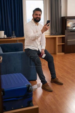 Photo for Pleased gentleman with travel documents seated on sofa arm holding mobile phone - Royalty Free Image