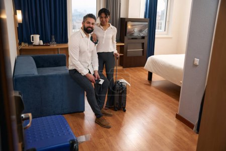 Téléchargez les photos : Homme souriant avec documents de voyage et sa compagne avec valise trolley dans la chambre d'hôtel - en image libre de droit