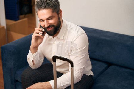 Téléchargez les photos : Homme d'affaires souriant avec bagages assis sur le canapé dans la chambre d'hôtel pendant la conversation téléphonique - en image libre de droit