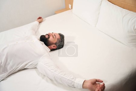 Téléchargez les photos : Touriste heureux souriant couché avec les yeux fermés sur le lit dans une suite de luxe - en image libre de droit