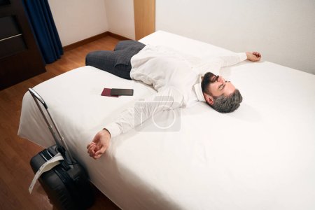 Téléchargez les photos : Souriant heureux homme élégant couché sur le lit dans sa chambre d'hôtel bien éclairée - en image libre de droit