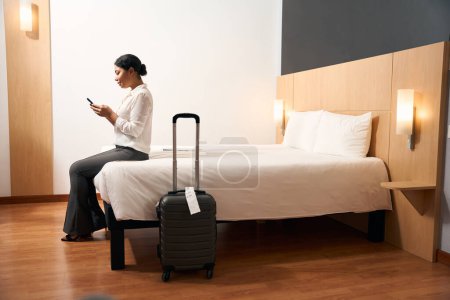 Téléchargez les photos : Vue latérale du touriste féminin concentré avec téléphone portable dans les mains assis sur le bord du lit dans la chambre d'hôtel - en image libre de droit