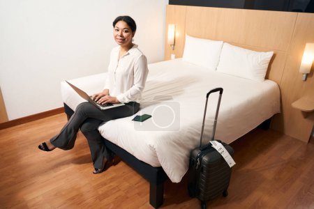 Photo for Smiling elegant young woman seated on edge of bed in hotel room typing on laptop - Royalty Free Image