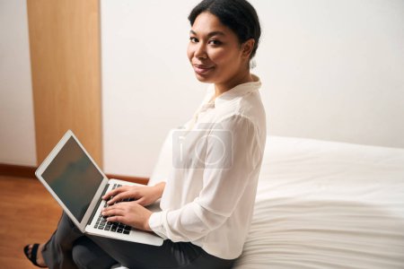 Photo for Contented stylish female entrepreneur with portable computer sitting on edge of bed in hotel room - Royalty Free Image