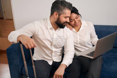 Photo for Pleased young man seated on sofa looking at laptop in hands of his smiling female companion - Royalty Free Image