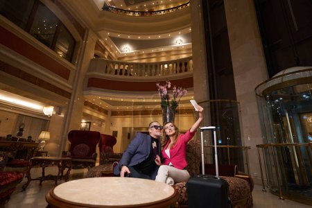 Téléchargez les photos : Couple voyageur prend un selfie dans la zone de loisirs de l'hôtel de luxe, autour de l'intérieur élégant - en image libre de droit
