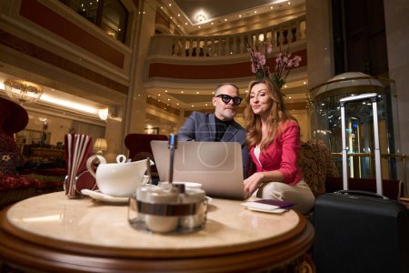Foto de Los cónyuges con un ordenador portátil se relajan en el vestíbulo de un hotel de lujo, el té se sirve en la zona de relajación - Imagen libre de derechos
